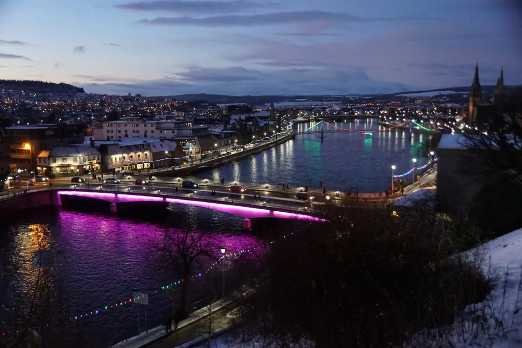 Inverness at dusk seen from above, with the River Ness, a bridge lit in magenta and the lights of the town centre