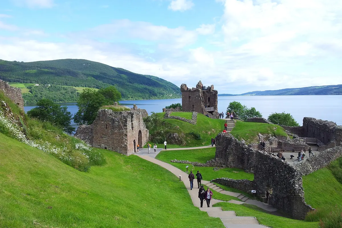 The ruins of Urquhart Castle on a green headland beside Loch Ness, with a path, visitors and hills behind